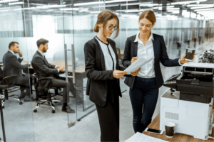 Two business women stand next to an office copier while looking at printed documents to show their importance.