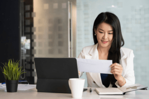 A businesswoman sits in front of her laptop at a desk reviewing office mail signifying an office postage meter