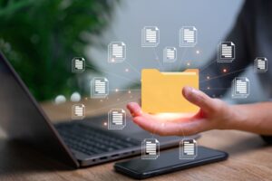 A close up of a hand hovering over a black laptop and black phone. The phone appears to be holding a file with offshoots of individual files, representing document management software.