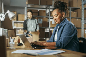 A smiling woman with curly hair working on a laptop in a warehouse office, with a postage meter on the desk beside her. In the background, a focused male colleague is preparing packages for shipping, surrounded by shelves filled with boxes.