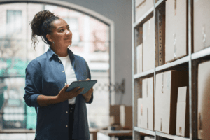 A woman in an office using a tablet while standing near shelves of packages, representing the efficiency and organization enabled by an office postage machine.