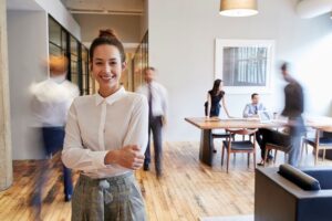 A smiling young professional is in the foreground as her coworkers work and walk behind her in a busy office signifying the need for a quality Copier Service Provider,