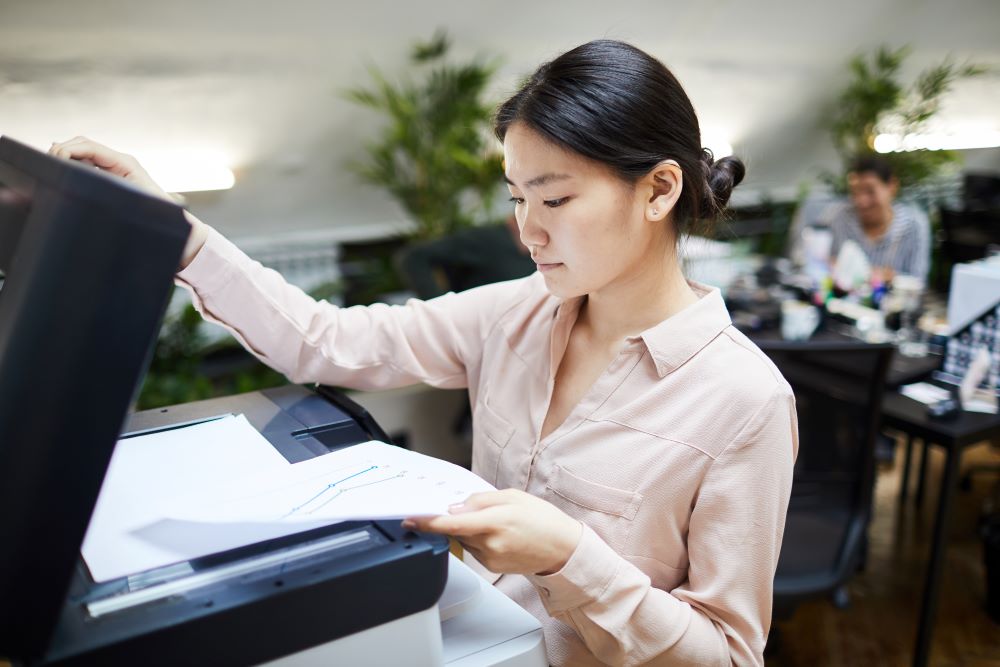 A young professional stands in front of a color office copier reviewing outputs while colleagues work behind her.