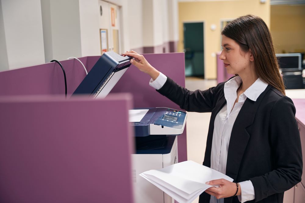 A young office worker stands in front of a large office copier holding outputs.