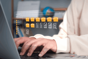 Close up of hands typing on a laptop in the foreground and a globe on a shelf in the background. Above the hands are icons within the file search feature, with folders and document icons on the screen, as a conceptual representation of digital records management.