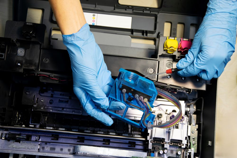 Close up of a copier service technician's hands, wearing blue gloves, holding a small screwdriver and interfacing with the interworking of a copier signifying copier maintenance.