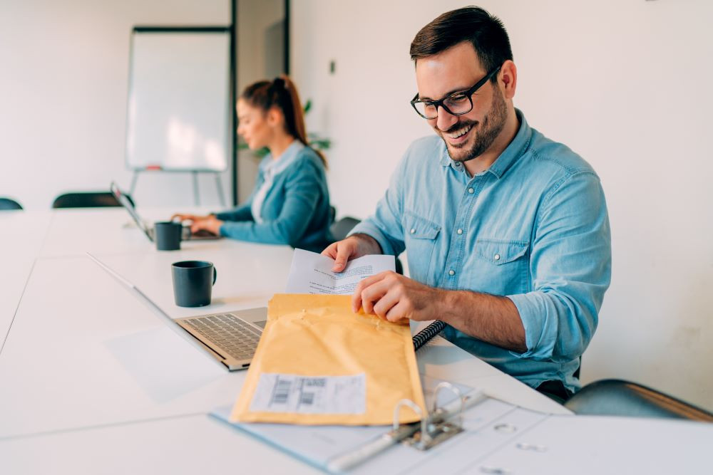 A smiling office worker puts papers into an envelope signifying the importance of a postage meter to keep business workflows moving forward.